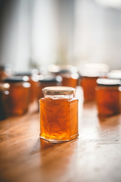A collection of hexagonal glass jars filled with amber-colored marmalade is arranged on a wooden surface, with soft light coming from a window in the background creating a warm and cozy atmosphere.