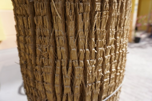 A close-up view of a textured surface made up of tightly packed, vertical strands of natural fibers. The fibers are light brown in color and appear to be tied together with small wire loops. The background is blurred, with hints of a pale yellow and gray environment.