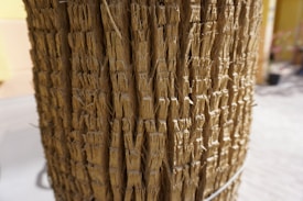 A close-up view of a textured surface made up of tightly packed, vertical strands of natural fibers. The fibers are light brown in color and appear to be tied together with small wire loops. The background is blurred, with hints of a pale yellow and gray environment.