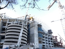 A large-scale construction project is underway, featuring a partially completed building with modern architectural elements. Tall cranes are positioned around the site, and scaffolding is visible on an unfinished structure. The foreground shows a crane vehicle, and the surrounding area is filled with metal beams and construction materials. Leafless trees frame the upper part of the image.