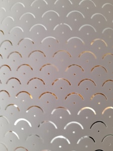 Metal surface with a repeating pattern of semi-circular cutouts and dots arranged in rows. The cutouts reflect light, creating subtle variations in color and texture.