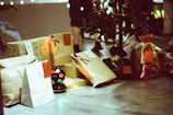 A collection of wrapped gifts is arranged on a floor beside a small Christmas tree, adorned with decorations. The scene includes several rectangular and bag-shaped presents, some with visible labels and embellishments. A colorful, round light fixture is placed among the gifts, adding a festive touch to the setting. The image captures a cozy, holiday atmosphere.