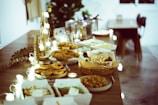 A beautifully arranged wooden table is laden with an assortment of foods, including baskets of bread, trays of pies, and plates of fried items. Soft, warm lighting is provided by string lights and candles, creating a cozy atmosphere. Incorporating natural and elegant elements, there is also some decorative foliage.