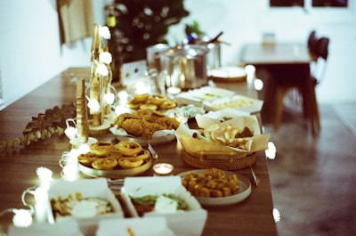 A beautifully arranged wooden table is laden with an assortment of foods, including baskets of bread, trays of pies, and plates of fried items. Soft, warm lighting is provided by string lights and candles, creating a cozy atmosphere. Incorporating natural and elegant elements, there is also some decorative foliage.