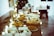 A beautifully arranged wooden table is laden with an assortment of foods, including baskets of bread, trays of pies, and plates of fried items. Soft, warm lighting is provided by string lights and candles, creating a cozy atmosphere. Incorporating natural and elegant elements, there is also some decorative foliage.