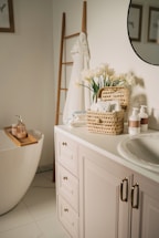 A serene and well-organized bathroom featuring a white bathtub with wooden accents, a countertop with a sink, and neatly arranged toiletries. There is a basket with flowers and towels on the counter, and a ladder-style towel rack beside the tub. The setting is bright and airy with framed artwork partially visible on the walls.