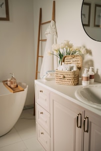 A serene and well-organized bathroom featuring a white bathtub with wooden accents, a countertop with a sink, and neatly arranged toiletries. There is a basket with flowers and towels on the counter, and a ladder-style towel rack beside the tub. The setting is bright and airy with framed artwork partially visible on the walls.