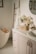 A serene and well-organized bathroom featuring a white bathtub with wooden accents, a countertop with a sink, and neatly arranged toiletries. There is a basket with flowers and towels on the counter, and a ladder-style towel rack beside the tub. The setting is bright and airy with framed artwork partially visible on the walls.