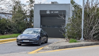 A modern electric car is parked in front of a gray building marked as a car wash. The car is positioned on a driveway bordered by yellow lines. The area is surrounded by greenery, and a lamp post is visible to the right.