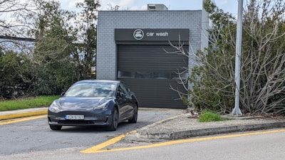 A modern electric car is parked in front of a gray building marked as a car wash. The car is positioned on a driveway bordered by yellow lines. The area is surrounded by greenery, and a lamp post is visible to the right.