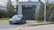 A modern electric car is parked in front of a gray building marked as a car wash. The car is positioned on a driveway bordered by yellow lines. The area is surrounded by greenery, and a lamp post is visible to the right.