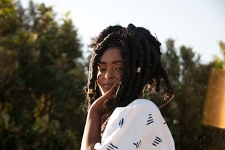 A person with long, textured hair styled in twists, wearing a white garment with black patterns, is positioned outdoors. The background is filled with lush green foliage against a bright blue sky.