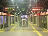 Interior of a modern train station platform with automated doors. Digital signs above display information in Japanese. An overhead clock shows the time as 18:56. Pillars with red and white signs are evenly spaced along the platform. A central structure with a fire extinguisher is positioned in the middle. The ambiance is calm and orderly with clear pathways marked by yellow tactile paving.