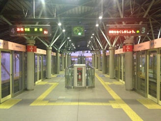 Interior of a modern train station platform with automated doors. Digital signs above display information in Japanese. An overhead clock shows the time as 18:56. Pillars with red and white signs are evenly spaced along the platform. A central structure with a fire extinguisher is positioned in the middle. The ambiance is calm and orderly with clear pathways marked by yellow tactile paving.