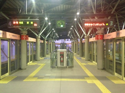 Interior of a modern train station platform with automated doors. Digital signs above display information in Japanese. An overhead clock shows the time as 18:56. Pillars with red and white signs are evenly spaced along the platform. A central structure with a fire extinguisher is positioned in the middle. The ambiance is calm and orderly with clear pathways marked by yellow tactile paving.