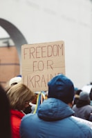 A group of people gathers at a protest, holding a cardboard sign with the words 'Freedom for Ukraine' written in red. People are dressed in winter clothing, and one person has a blue coat.