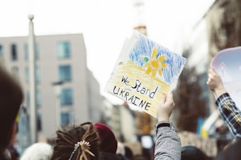 A gathering of people with signs expressing support for Ukraine. One prominently displayed sign features a drawing of a sunflower with the colors blue and yellow, alongside the words 'We Stand with Ukraine'. The setting appears to be an urban environment, with blurred buildings in the background.