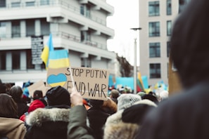 A large group of people is gathered in a city setting, holding signs and banners advocating for peace. Prominent among them is a sign with the message 'Stop Putin, End War' along with the blue and yellow colors of the Ukrainian flag. The crowd appears diverse, wearing winter clothing, and is located in a modern urban area with multi-story buildings visible in the background.
