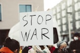 A group of people are participating in a protest, holding a sign with the words 'Stop War' written on it. The setting appears to be outdoors in an urban environment, with buildings in the background.