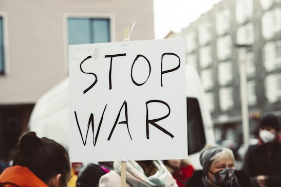 A group of people are participating in a protest, holding a sign with the words 'Stop War' written on it. The setting appears to be outdoors in an urban environment, with buildings in the background.