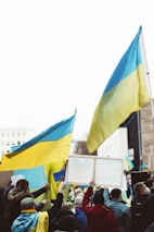 A group of people holding Ukrainian flags and signs with messages of peace, gathered in a public space. The atmosphere is lively with many participants wearing yellow and blue clothing. The crowd is diverse, consisting of individuals wrapped in flags and interacting with one another.