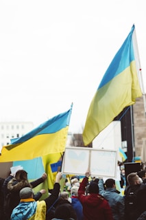 A group of people holding Ukrainian flags and signs with messages of peace, gathered in a public space. The atmosphere is lively with many participants wearing yellow and blue clothing. The crowd is diverse, consisting of individuals wrapped in flags and interacting with one another.