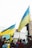 A group of people holding Ukrainian flags and signs with messages of peace, gathered in a public space. The atmosphere is lively with many participants wearing yellow and blue clothing. The crowd is diverse, consisting of individuals wrapped in flags and interacting with one another.