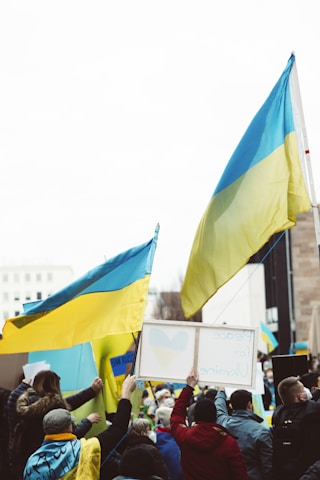 A group of people holding Ukrainian flags and signs with messages of peace, gathered in a public space. The atmosphere is lively with many participants wearing yellow and blue clothing. The crowd is diverse, consisting of individuals wrapped in flags and interacting with one another.