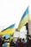 A group of people holding Ukrainian flags and signs with messages of peace, gathered in a public space. The atmosphere is lively with many participants wearing yellow and blue clothing. The crowd is diverse, consisting of individuals wrapped in flags and interacting with one another.