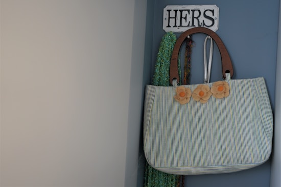 A striped bag hangs on a wall hook labeled 'HERS.' The bag has decorative flowers attached to it. The wall is painted in blue, and a scarf can be seen behind the bag.