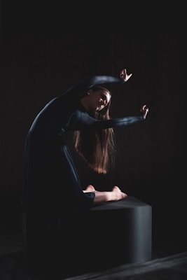 A person with long hair is gracefully posed in a dark space, sitting on a low platform with arms extended above and around the head. The lighting emphasizes the contours of the body, creating a dramatic and artistic effect.