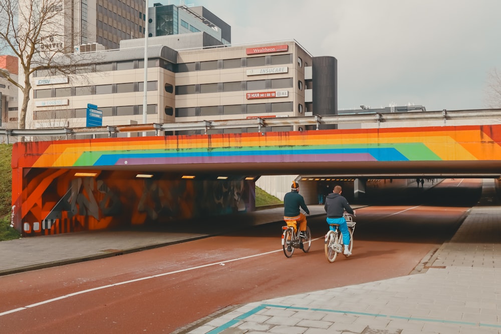 a couple of people riding bikes under a rainbow colored bridge