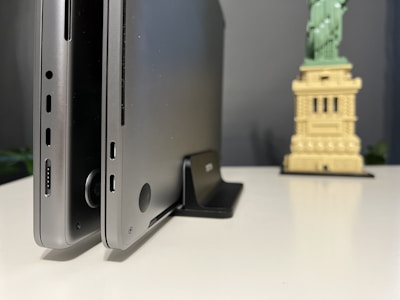 Two laptops in a vertical standing position are placed on a desk against a neutral background. In the foreground, the edges and ports of the laptops are visible, suggesting a focus on design and technology. In the background, there is a small model resembling the Statue of Liberty, contributing an element of decor or personal touch to the workspace.