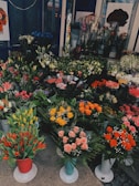 A colorful display of various flowers arranged in pots and bouquets. The assortment includes tulips, roses, lilies, and other florals. The flowers are arranged in different colors like red, yellow, pink, and orange. The shop display is vibrant and abundant, providing a lush and inviting atmosphere.