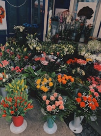 A colorful display of various flowers arranged in pots and bouquets. The assortment includes tulips, roses, lilies, and other florals. The flowers are arranged in different colors like red, yellow, pink, and orange. The shop display is vibrant and abundant, providing a lush and inviting atmosphere.
