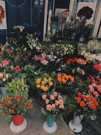 A colorful display of various flowers arranged in pots and bouquets. The assortment includes tulips, roses, lilies, and other florals. The flowers are arranged in different colors like red, yellow, pink, and orange. The shop display is vibrant and abundant, providing a lush and inviting atmosphere.