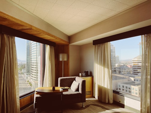 A cozy corner of a hotel room with large windows offering a view of modern skyscrapers. A comfortable armchair is positioned next to a small round table. Soft sunlight streams through sheer curtains, casting gentle shadows. A lamp and a small cabinet with a coffee maker are also visible.
