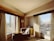 A cozy corner of a hotel room with large windows offering a view of modern skyscrapers. A comfortable armchair is positioned next to a small round table. Soft sunlight streams through sheer curtains, casting gentle shadows. A lamp and a small cabinet with a coffee maker are also visible.