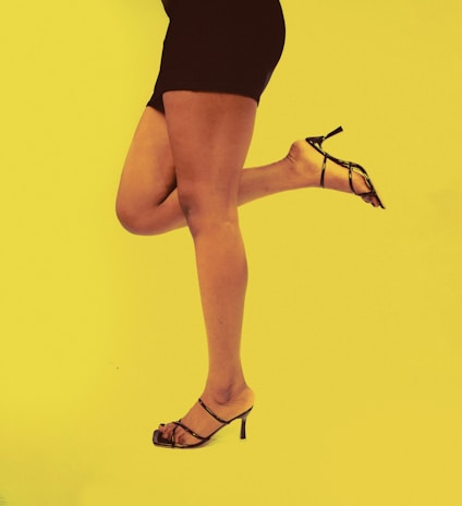 A person wearing a black dress and high-heeled strappy sandals is posing against a bright yellow background. The person is lifting one leg, highlighting the elegance of the footwear.
