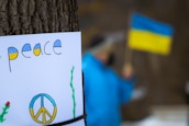 A poster attached to a tree features the word 'peace' and a peace sign painted in the colors of the Ukrainian flag. In the blurred background, a person wearing blue is holding a Ukrainian flag.