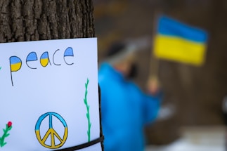 A poster attached to a tree features the word 'peace' and a peace sign painted in the colors of the Ukrainian flag. In the blurred background, a person wearing blue is holding a Ukrainian flag.
