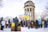 A group of people gathered in a snowy area, holding Ukrainian and Canadian flags. They stand in front of a historic clock tower with columns. Most individuals are wearing winter clothing, including jackets and hats. There are bare trees in the background, indicating a winter season.