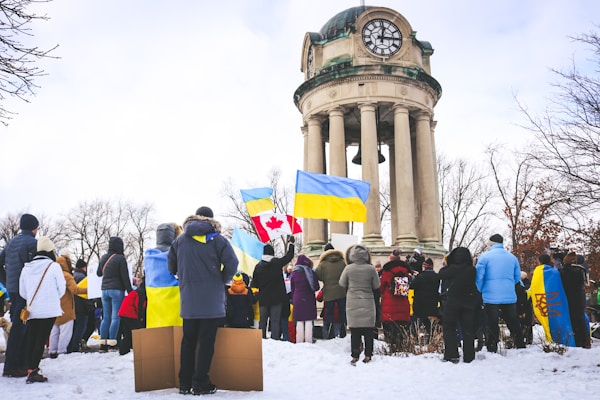 A group of people gathered in a snowy area, holding Ukrainian and Canadian flags. They stand in front of a historic clock tower with columns. Most individuals are wearing winter clothing, including jackets and hats. There are bare trees in the background, indicating a winter season.