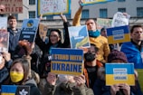 A group of people are holding various signs in support of Ukraine, with messages like 'I Stand with Ukraine' and 'Stop Putin'. Many individuals wear yellow and blue, the colors of the Ukrainian flag. The expressions on their faces convey determination and solidarity.