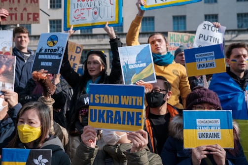 A group of people are holding various signs in support of Ukraine, with messages like 'I Stand with Ukraine' and 'Stop Putin'. Many individuals wear yellow and blue, the colors of the Ukrainian flag. The expressions on their faces convey determination and solidarity.