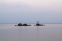 A serene seascape with a calm, glassy water surface. A small rocky island is situated in the center with a traditional red torii gate standing prominently on top. The sky is overcast with soft, muted hues of gray and blue, creating a peaceful atmosphere.