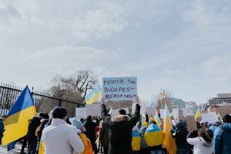 A group of people are gathered outside, holding various signs and Ukrainian flags. The scene suggests a protest or demonstration, with participants bundled in warm clothing. The central sign reads 'Honor the Budapest Memorandum'. Buildings and trees are visible in the background.