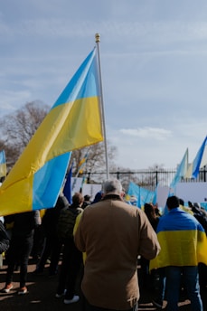 A crowd of people holding blue and yellow flags, which appear to be Ukrainian flags, gathered possibly for a protest or rally. The scene is outdoors with a clear sky and some leafless trees in the background.