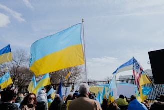 A crowd gathers outdoors holding Ukrainian flags, with one large flag prominently displayed in the foreground. An American flag is also visible among the group. The event takes place under a partly cloudy sky, and the background includes a black metal fence and leafless trees, suggesting a colder season.