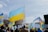 A crowd gathers outdoors holding Ukrainian flags, with one large flag prominently displayed in the foreground. An American flag is also visible among the group. The event takes place under a partly cloudy sky, and the background includes a black metal fence and leafless trees, suggesting a colder season.